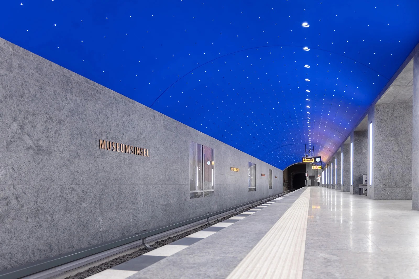 Image (Station entrance detail with escalators): "Museumsinsel U-Bahn station entrance at blue hour showing contemporary design without traditional Berlin underground light portal, open granite parapet with WE-EF VLR100 linear wall luminaires integrated into transparent covered wall boxes providing asymmetric wall wash downlighting, glass-enclosed escalators leading to 180-meter platform beneath Spree Canal, iconic blue U-Bahn signage, designed by Max Dudler Architects as underground entrée to Museum Island cultural institutions completing U5 line connection between Brandenburger Tor and Alexanderplatz