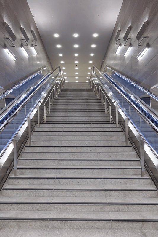 Unter den Linden station escalator corridor showing three-level vertical circulation from 14-meter-deep U5 platform to surface where U6 line crosses above, WE-EF DAC240 LED surface-mounted luminaires with integrated electronic control gear mounted on lift systems for maintenance access above escalators, identical appearance and 3000K warm white lighting technology creating uniform calm restful atmosphere designed by High Light Berlin lighting planner Thomas Spreen