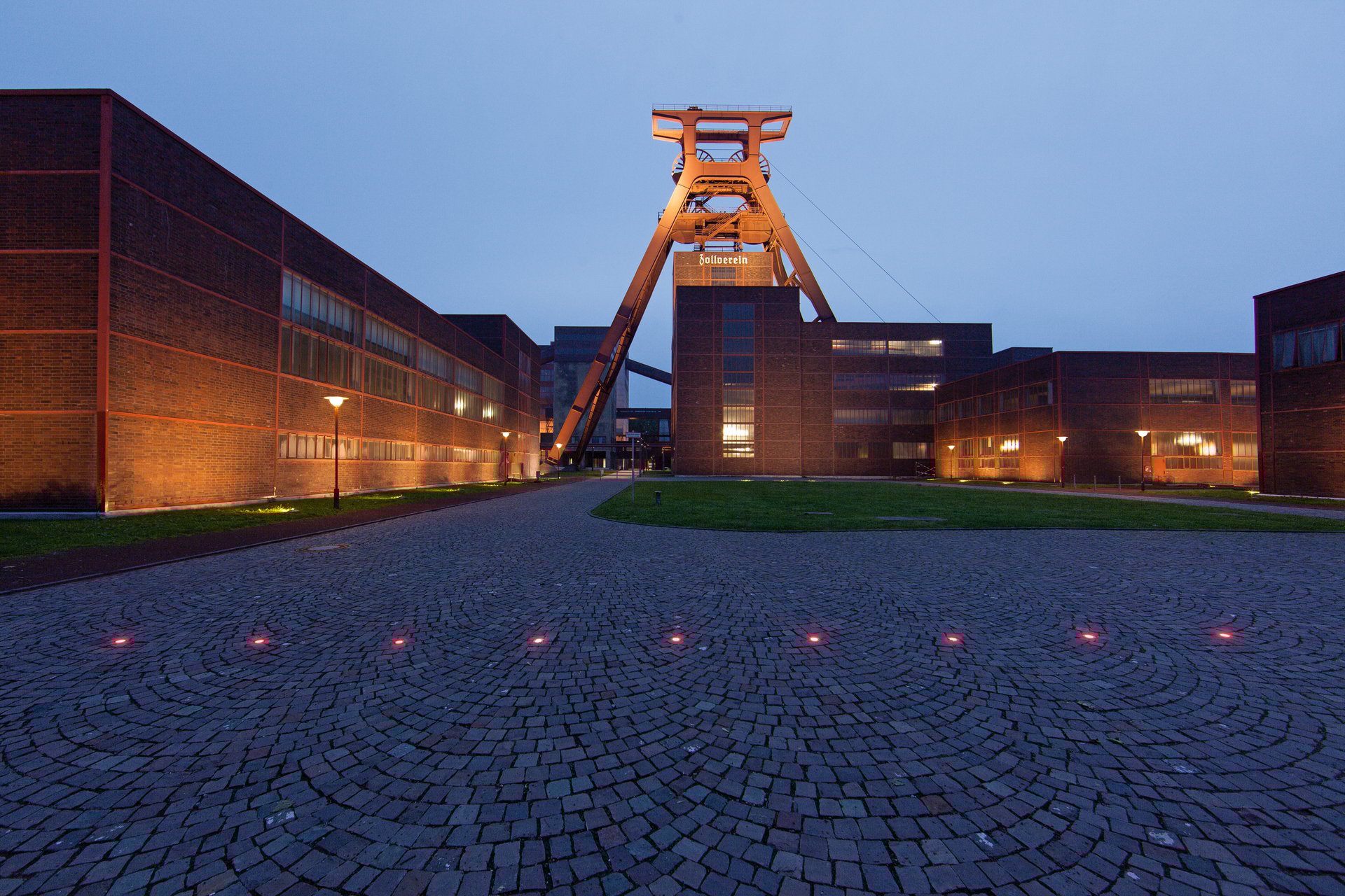 Frontal dusk view of Zeche Zollverein in Essen with the central headframe, warmly lit industrial buildings on either side, and red WE-EF ETC119 inground uplights set into the circular cobblestone pattern of the forecourt