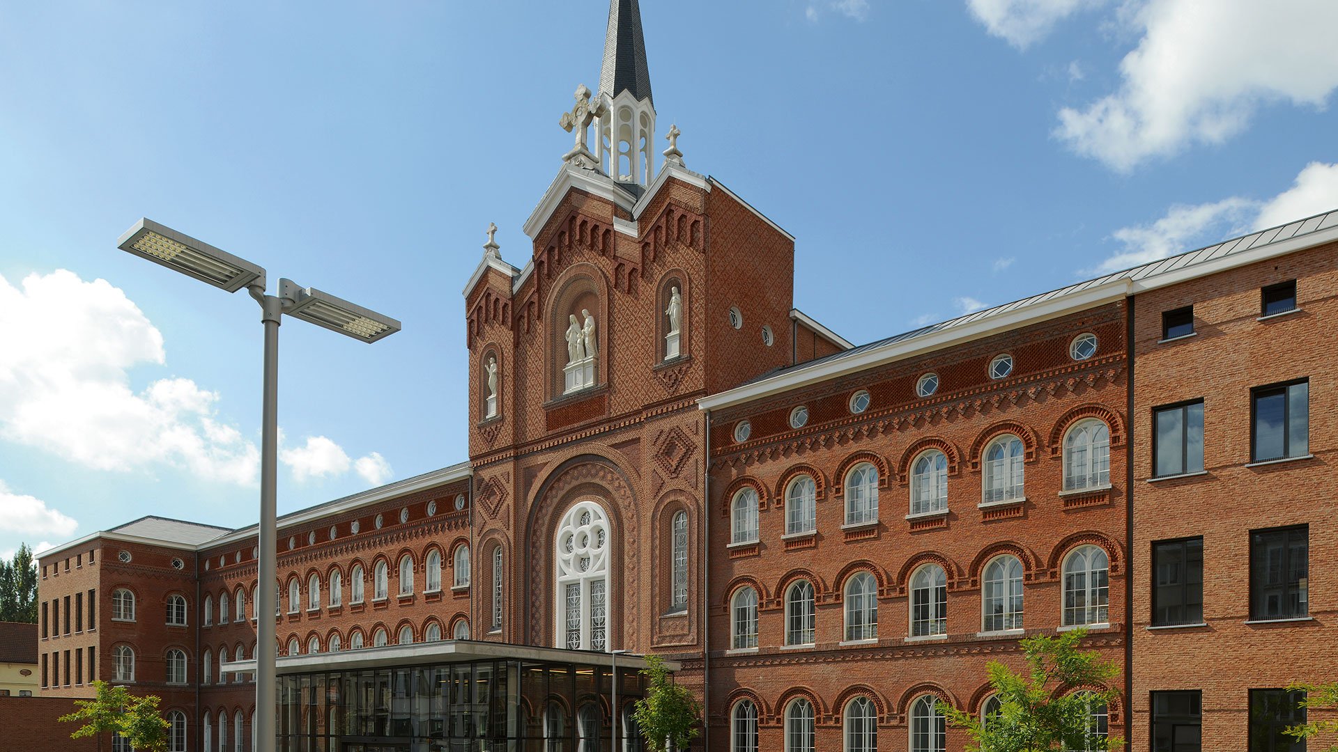 VFL540 LED street luminaire on a single pole in the foreground, with the ornate red brick facade and Gothic chapel tower of St. Vincentius Hospital Antwerp in the background under a blue sky