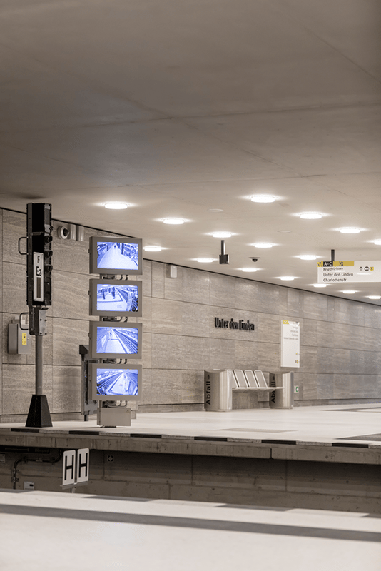 Unter den Linden U-Bahn platform level showing shell limestone wall cladding with digital information displays, 410 WE-EF LED recessed ceiling luminaires providing uniform soft 3000K warm white illumination meeting strict transport infrastructure standards, project-specific DOC240 luminaires with modified mounting elements optimized for installation cavities created years earlier during cut-and-cover construction, completed 2020 for BVG Berliner Verkehrsbetriebe completing prestigious U5 line extension