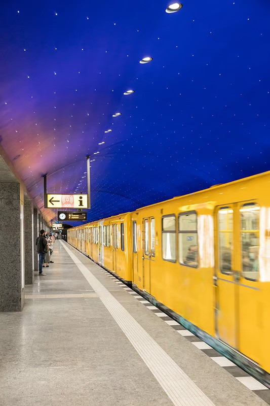 Museumsinsel U-Bahn platform showing 180-meter-long arched tunnel with ultramarine blue starry sky ceiling featuring 6,662 illuminated points, continuous row of WE-EF DOC220 recessed downlights with 3000K color temperature mounted above platform edge providing standards-compliant functional lighting tested and approved by Berliner Verkehrsbetriebe (BVG) for optimal glare protection for passengers and train drivers, station located beneath Spree Canal south of Schlossbrücke serving as underground entrance to Museum Island cultural institutions