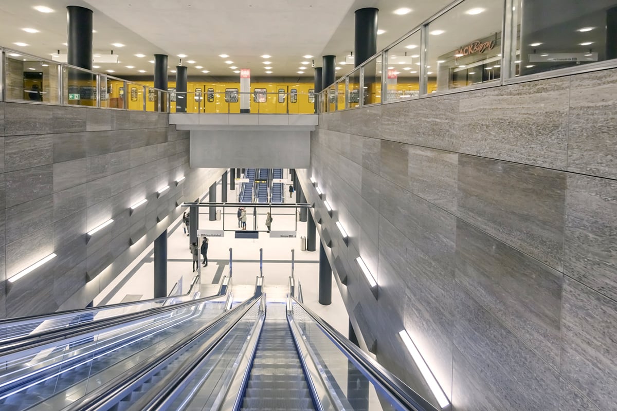 Dramatic vertical view through Unter den Linden U-Bahn station showing spacious prestigious architecture with wide opening connecting three levels from U5 platform 14 meters below to U6 crossing near surface, shell limestone walls creating timelessly elegant character, comprehensive WE-EF lighting system including 410 DOC240 recessed ceiling luminaires with custom acrylic covers bolted and wire-cable-secured against vibration, DAC240 surface-mounted luminaires on maintenance lifts, yellow U6 train visible at upper level, designed by Ingrid Hentschel and Prof. Axel Oestreich emphasizing principle that users make station lively